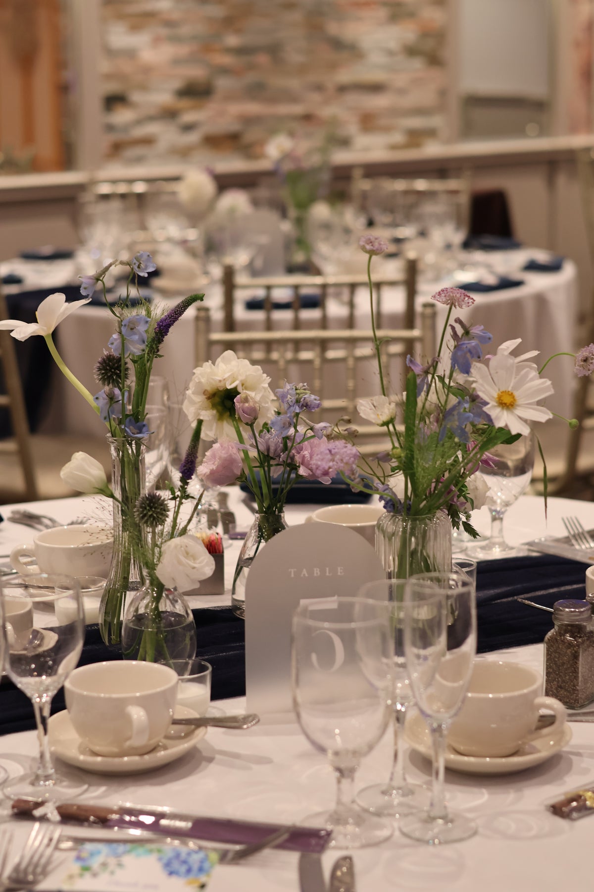A styled event table setting with white tablecloths, white teacups and saucers, clear wine glasses, silverware, and multiple floral centerpieces in glass vases. The background shows more tables and chairs in a banquet hall with a brick wall.
